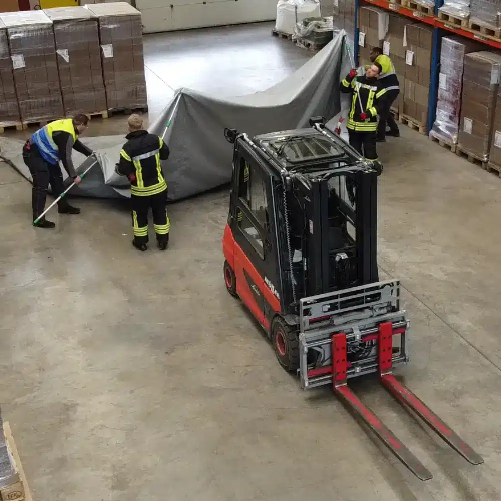 Einsatz der Brandbegrenzungsdecke an einem E-Gabelstapler in einer Lagerhalle durch Feuerwehr.